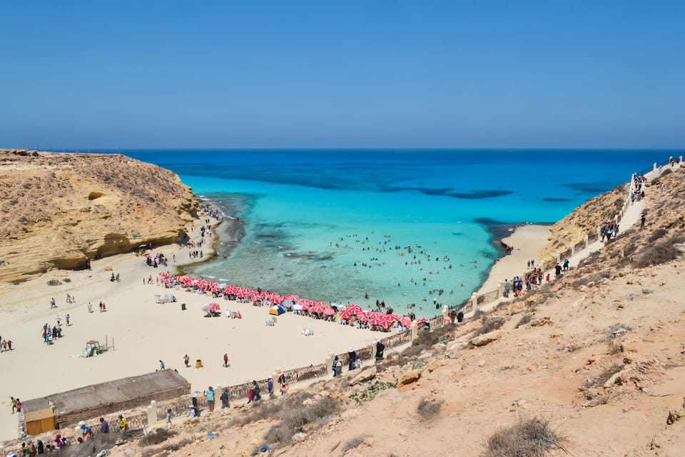 Una playa costera con aguas turquesas cristalinas, una orilla arenosa y acantilados rocosos.