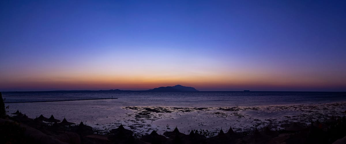 A beach at sunset with a pier extending into the water and a distant mountain.