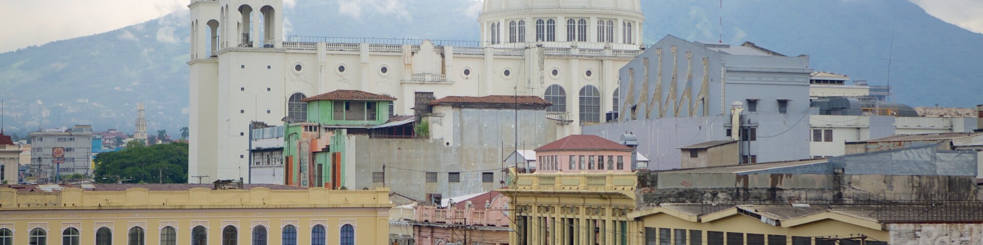 Paisagem urbana histórica com uma catedral branca imponente, apresentando uma cúpula grande, um campanário e montanhas ao fundo.