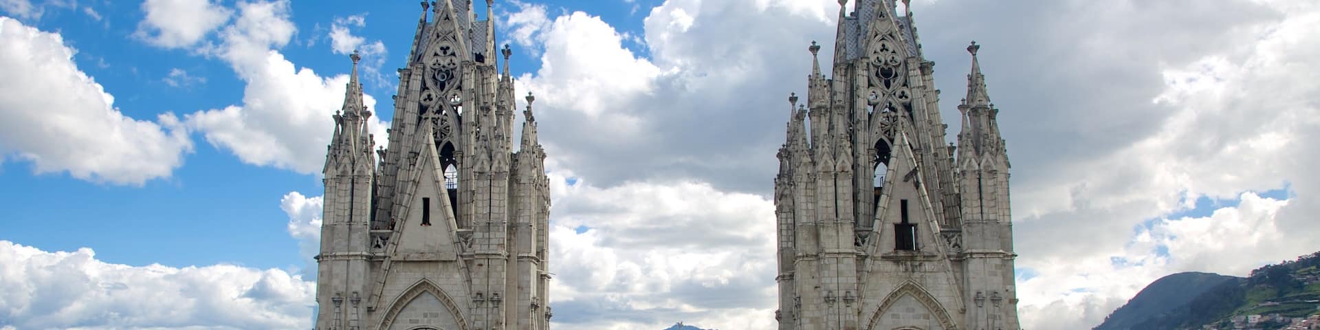 Two ornate Gothic towers with large clocks, set against a backdrop of a city and mountains under a partly cloudy sky.