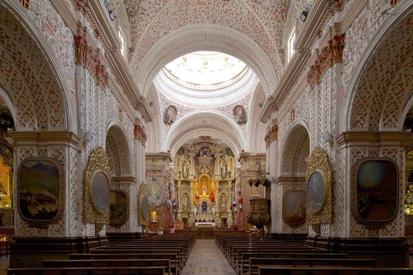 L’intérieur d’une église richement décorée, avec des motifs complexes sur le plafond, un autel central et des rangées de bancs.