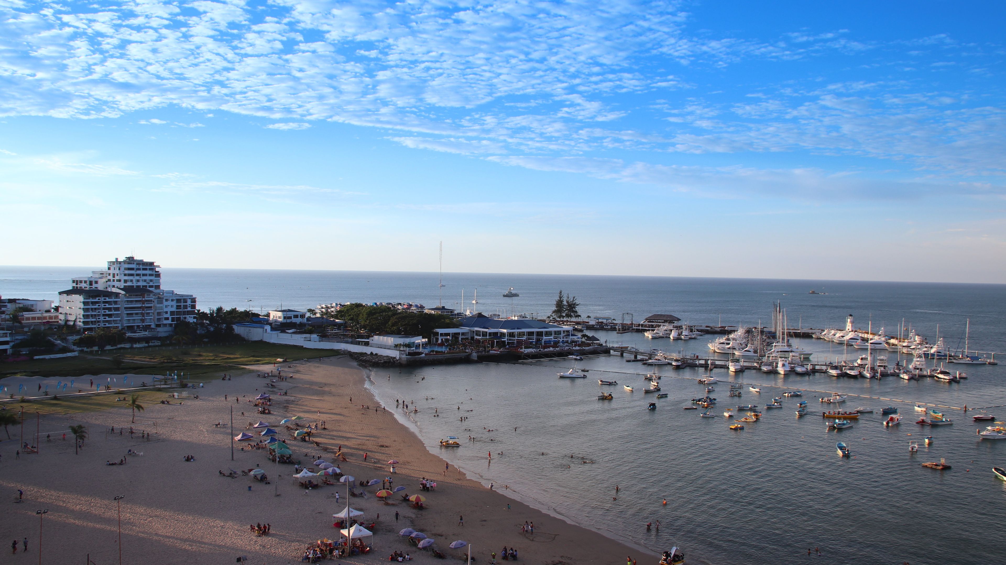 A coastal scene with a beach, a marina with boats, and a building complex.