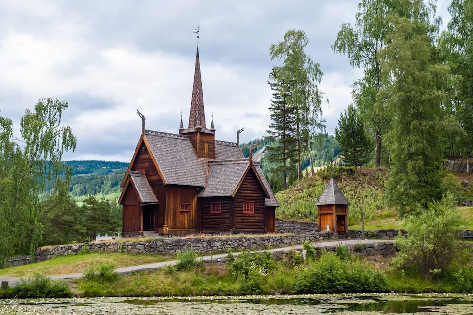 Une église en bois, dotée d’un clocher imposant, entourée d’une végétation luxuriante et bordée d’un étang où flottent des nénuphars.