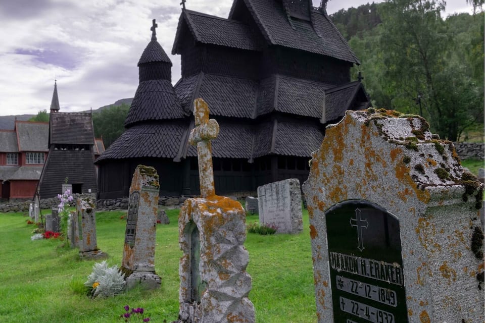 Eine Kirche mit einem steilen, dunklen Dach und mehreren Türmen, umgeben von Grabsteinen auf einem grasbewachsenen Friedhof.