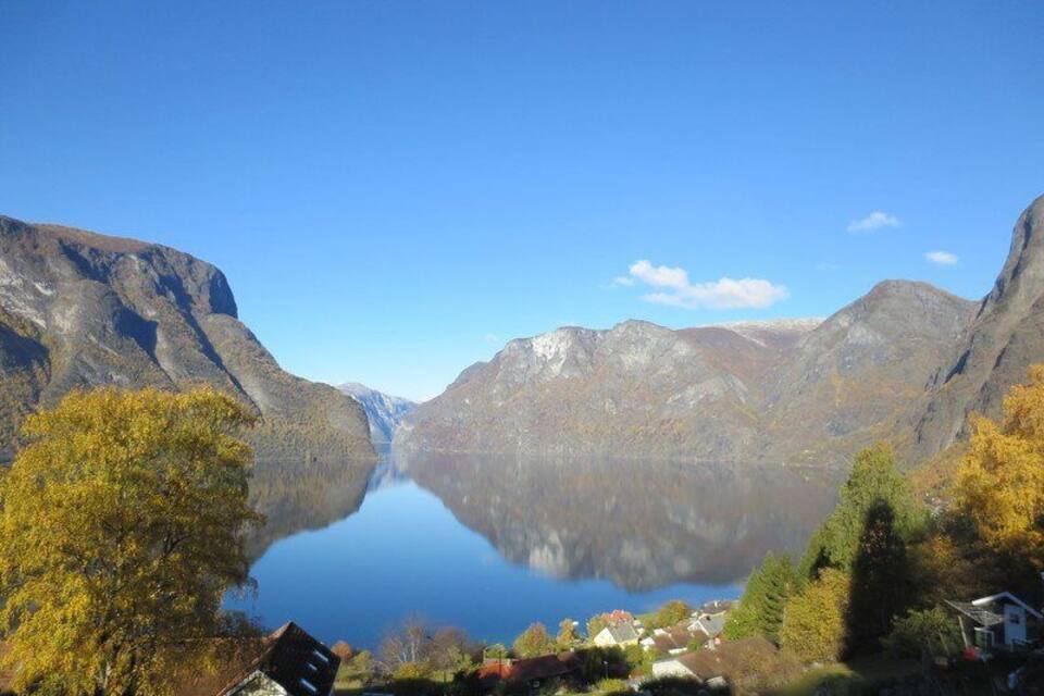 Un llac tranquil envoltat de muntanyes, amb arbres de tons tardans i una casa petita al primer pla.