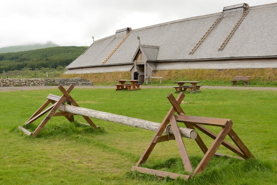 A wooden structure with picnic tables in a grassy field, a stone wall, and a building with a sloped roof in the background.