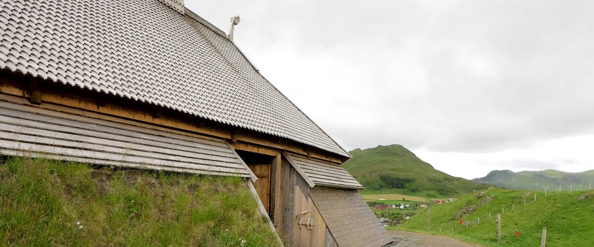 A traditional wooden house with a sloped roof, set in a rural landscape with green hills in the background.