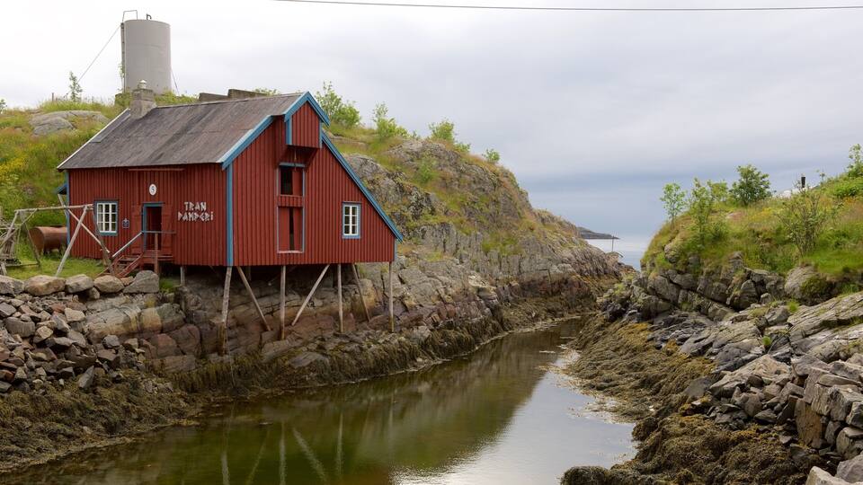 A red wooden house with the name "Tran Nampadi" on its side, situated by a rocky shoreline with a calm body of water in the background.