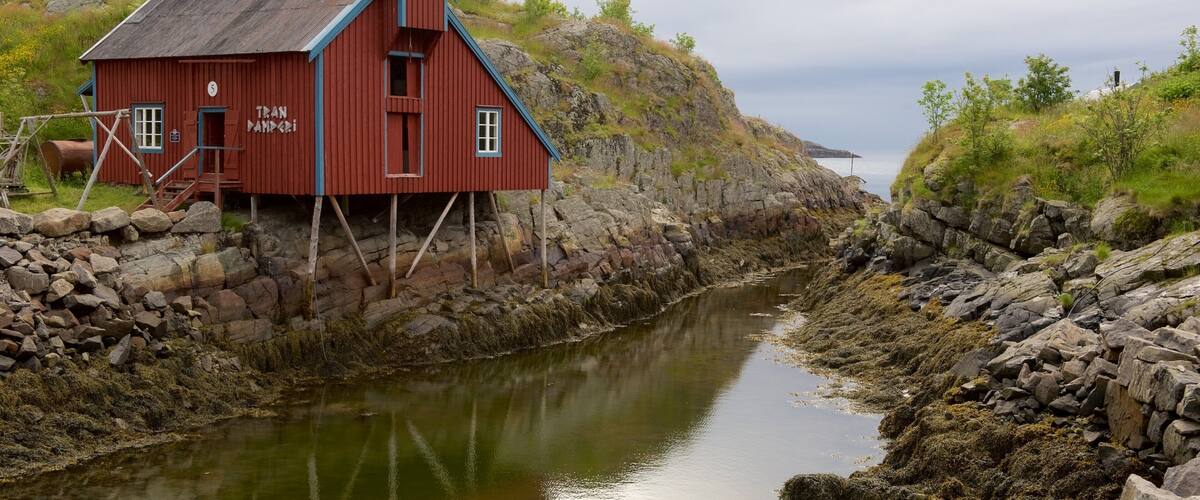 A red wooden house with the name "Tran Nampadi" on its side, situated by a rocky shoreline with a calm body of water in the background.