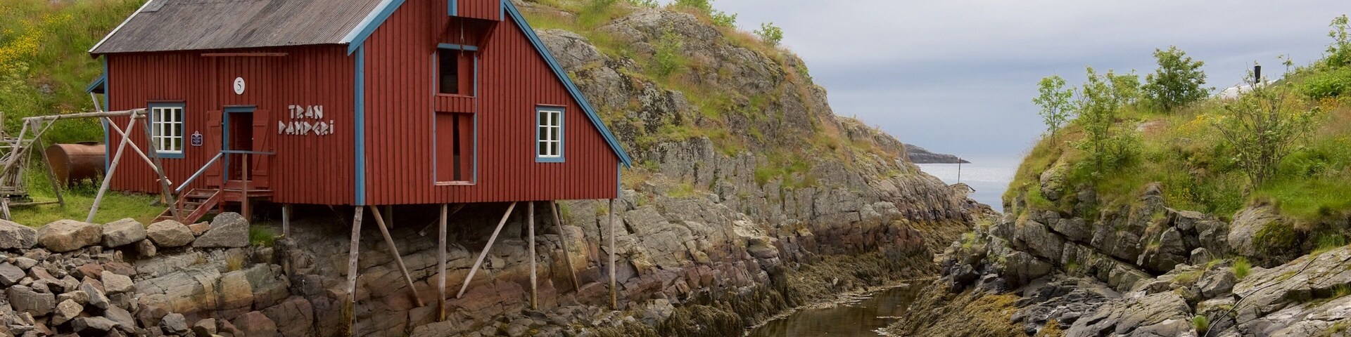 A red wooden house with the name "Tran Nampadi" on its side, situated by a rocky shoreline with a calm body of water in the background.