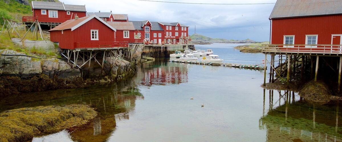 A coastal village with red buildings on stilts over water, a dock with boats, and a mountainous backdrop.