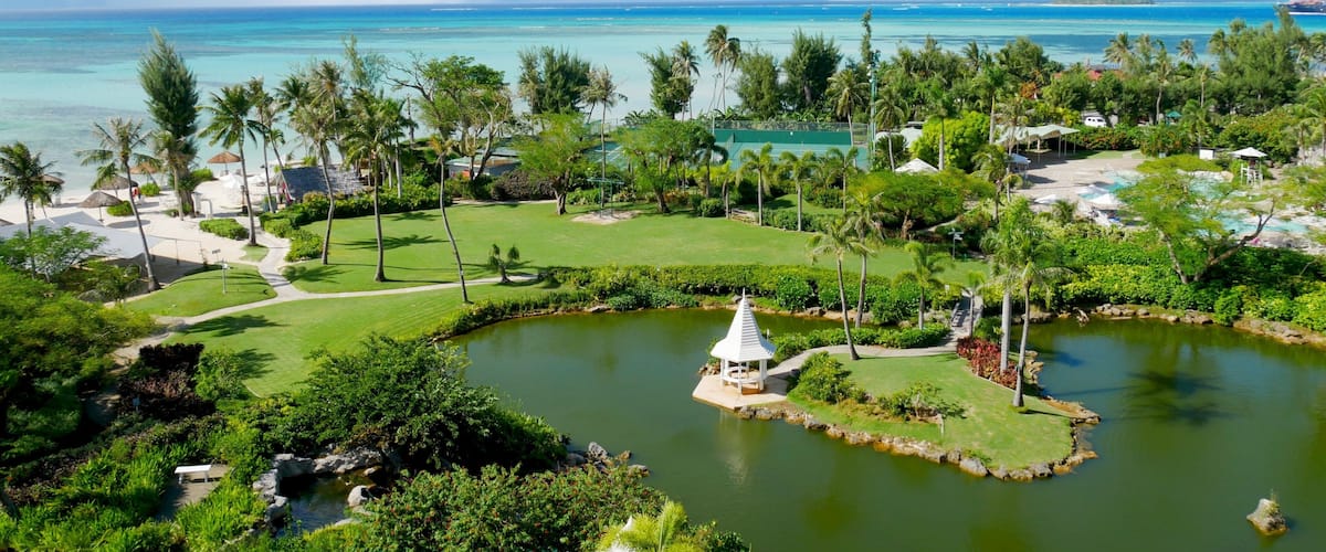A tropical resort with a golf course, a pond with a gazebo, and a beach with palm trees.