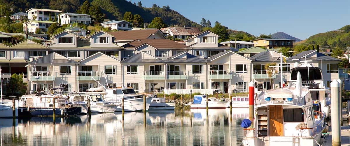 A marina with boats docked in calm water, surrounded by multi-story houses with balconies, set against a backdrop of green hills.
