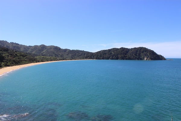 Eine Küstenlandschaft mit einem Sandstrand, klarem türkisblauem Wasser und einem bewaldeten Hügel im Hintergrund.
