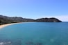 A coastal landscape with a sandy beach, clear turquoise waters, and a forested hill in the background.