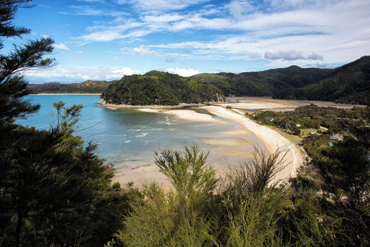 A coastal landscape with a sandy beach, clear blue waters, and a forested hill in the background.