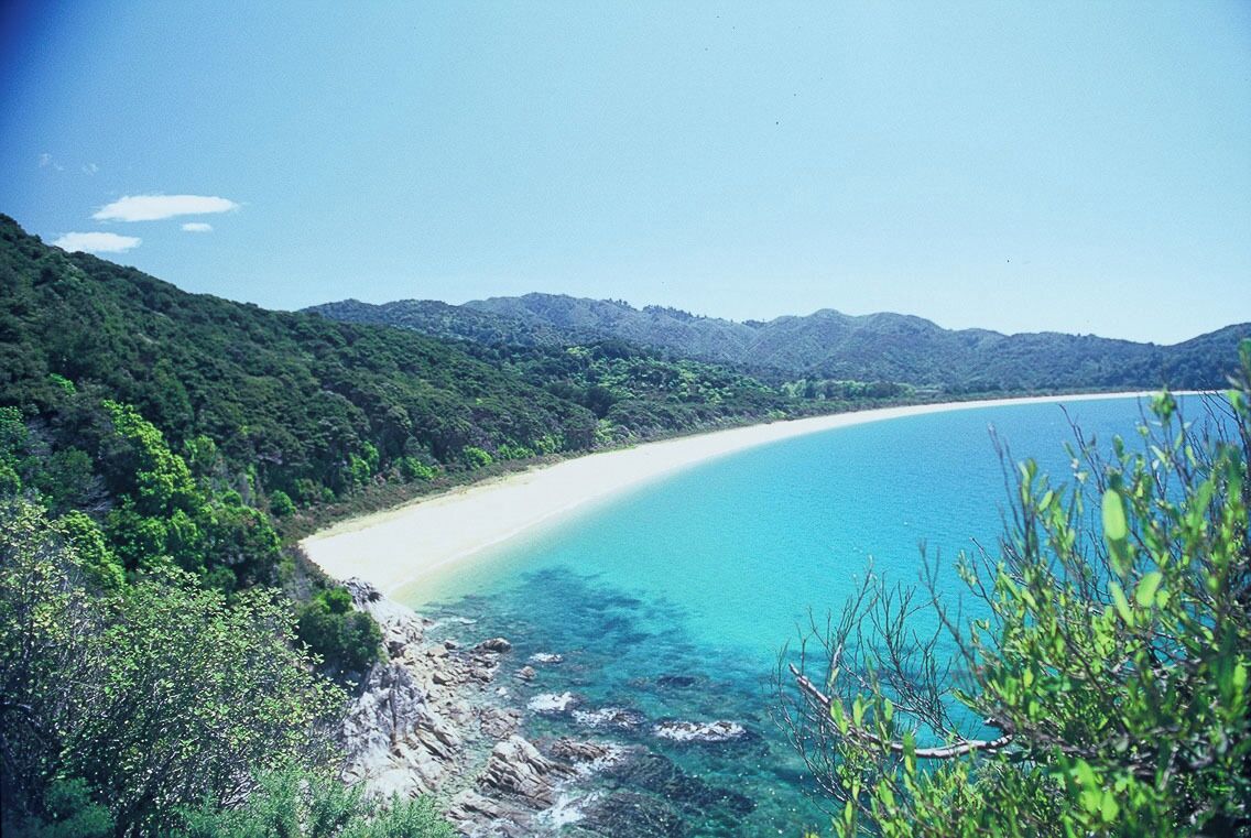 Un paisaje costero con mar azul cristalino, una playa arenosa y colinas verdes exuberantes.