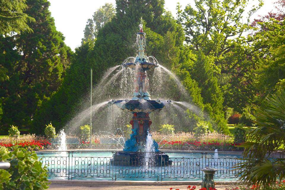 A fountain with water jets in a park with trees and flowers.