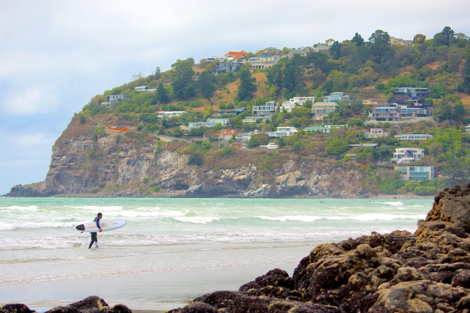 Una playa con olas, un surfista y casas en una colina.