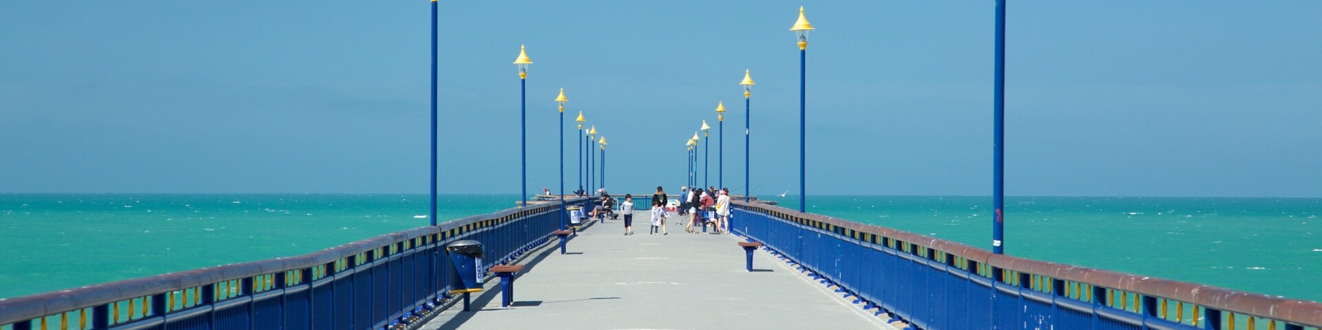 Eine Steg mit blauen Geländern und gelben Straßenlaternen, der zum Meer führt.