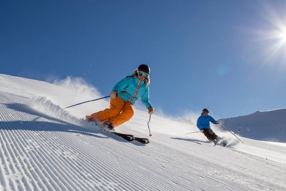 Two skiers on a snowy slope with a clear blue sky and a bright sun in the background.