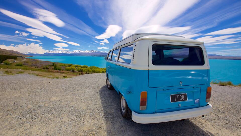 A blue and white van parked on a gravel road with a scenic view of a lake and mountains in the background.