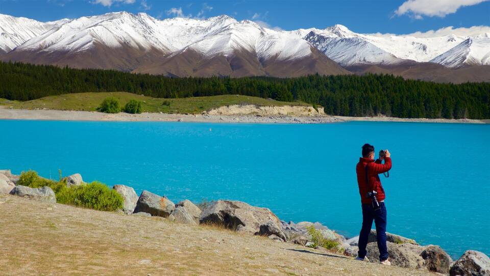 A person taking a photo of a lake with mountains in the background.