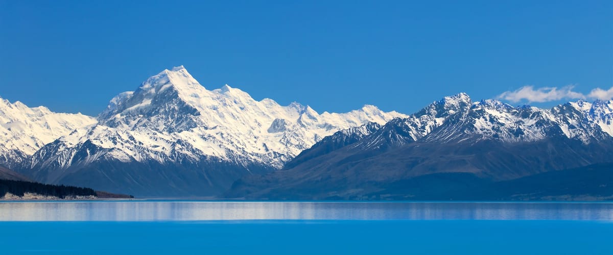 A mountain range with snow-capped peaks and a calm blue lake in the foreground.