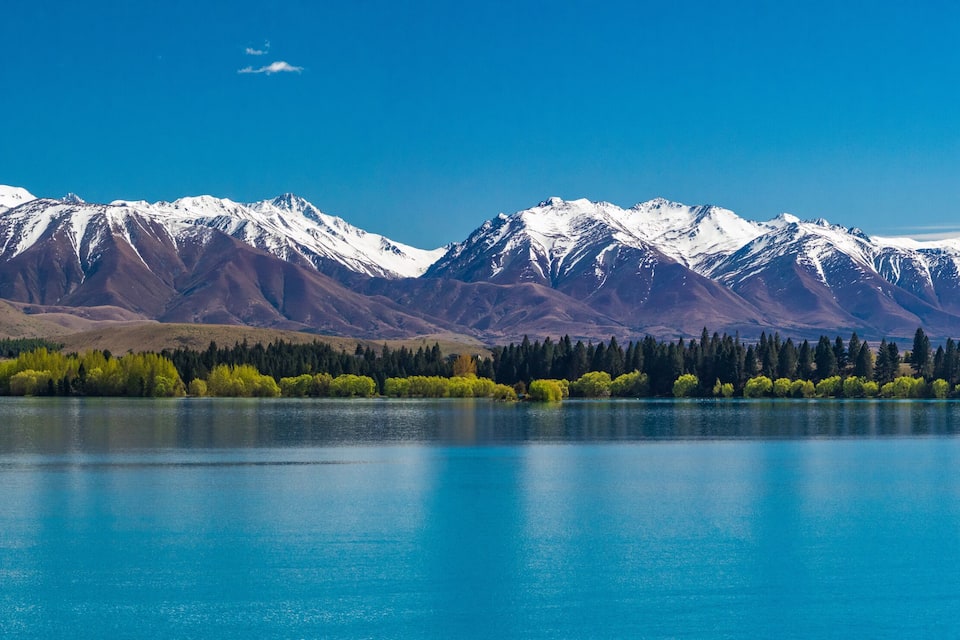 A mountain range with snow-capped peaks, a calm lake, and a forested shoreline.