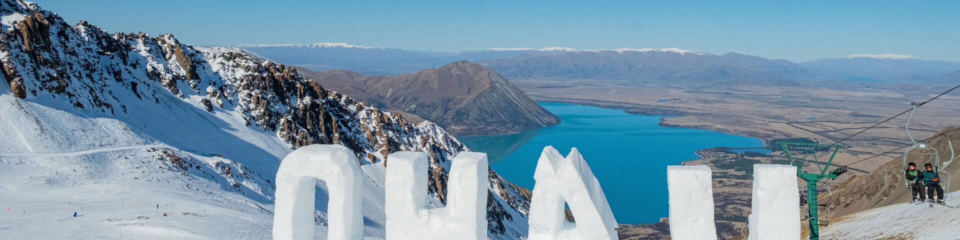 A snowy mountain landscape with the word "OHALU" carved in ice, a clear blue lake, and a ski lift in the background.