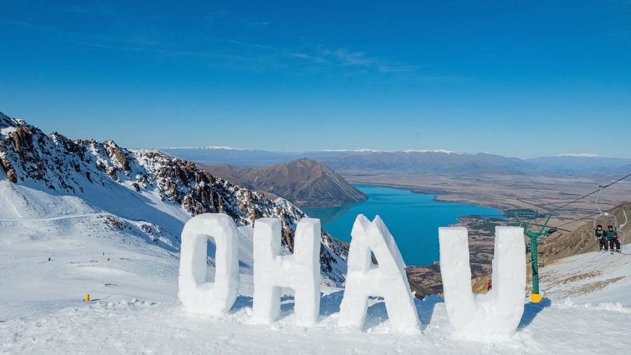 Paisaje nevado de montaña con la palabra "OHALU" tallada en hielo, un lago azul cristalino y un telesilla al fondo.