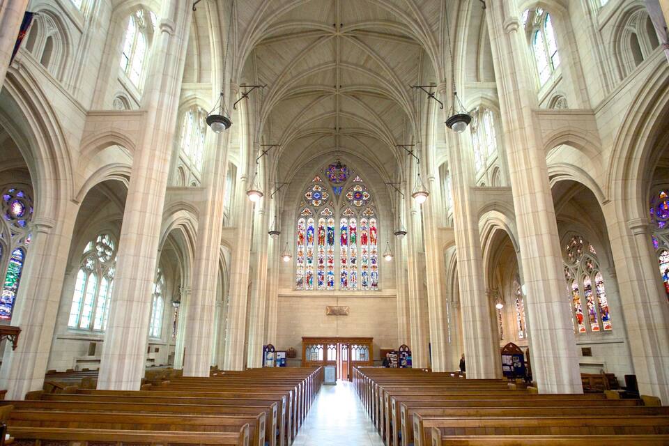 A cathedral interior with high vaulted ceilings, stained glass windows, and rows of wooden pews.