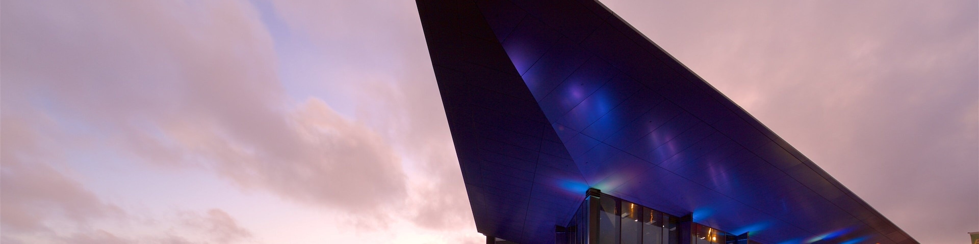 Un edificio moderno con un gran techo triangular y fachada de vidrio, frente a un cielo al atardecer.