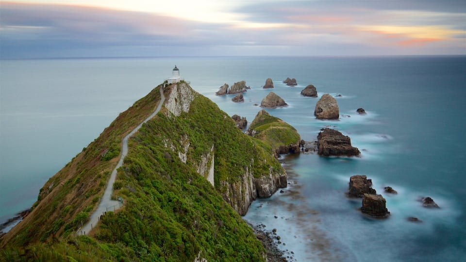 A coastal landscape with a lighthouse on a green hill, a winding path, and rocky outcrops in the sea.
