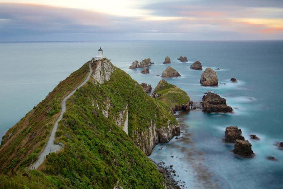 A coastal landscape with a lighthouse on a green hill, a winding path, and rocky outcrops in the sea.