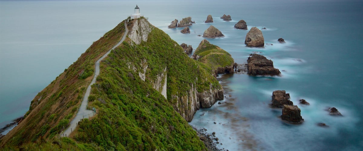 A coastal landscape with a lighthouse on a green hill, a winding path, and rocky outcrops in the sea.