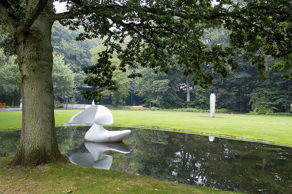Ein Park mit Teich, einem großen Baum und einer weißen Skulptur.
