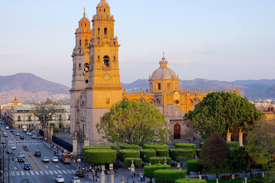 A historic cathedral with twin towers and a dome, surrounded by a well-maintained garden and a busy street.