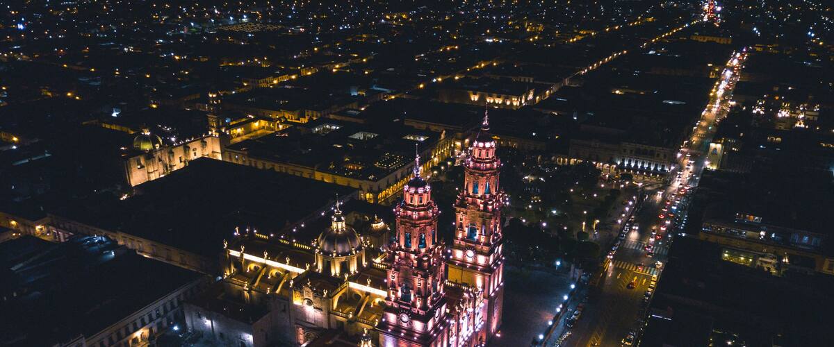 An illuminated cathedral with a prominent tower, surrounded by a cityscape at night.