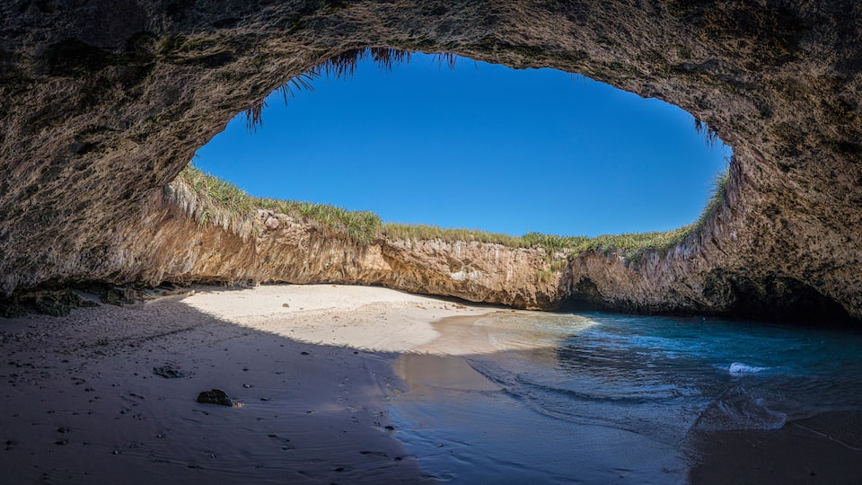 A natural cave opening to a beach with clear blue water and sandy shore.