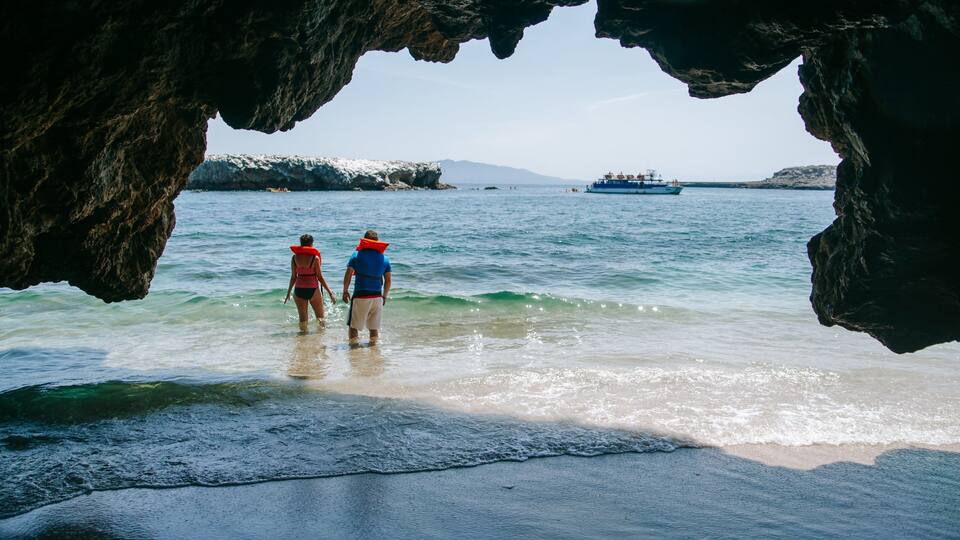 Two people walking in shallow water near a rocky cave entrance, with a large boat in the distance.