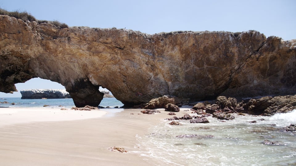 A rocky arch over a sandy beach with clear blue water.