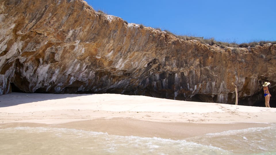 A beach with a large rock formation and a person in a swimsuit and hat.