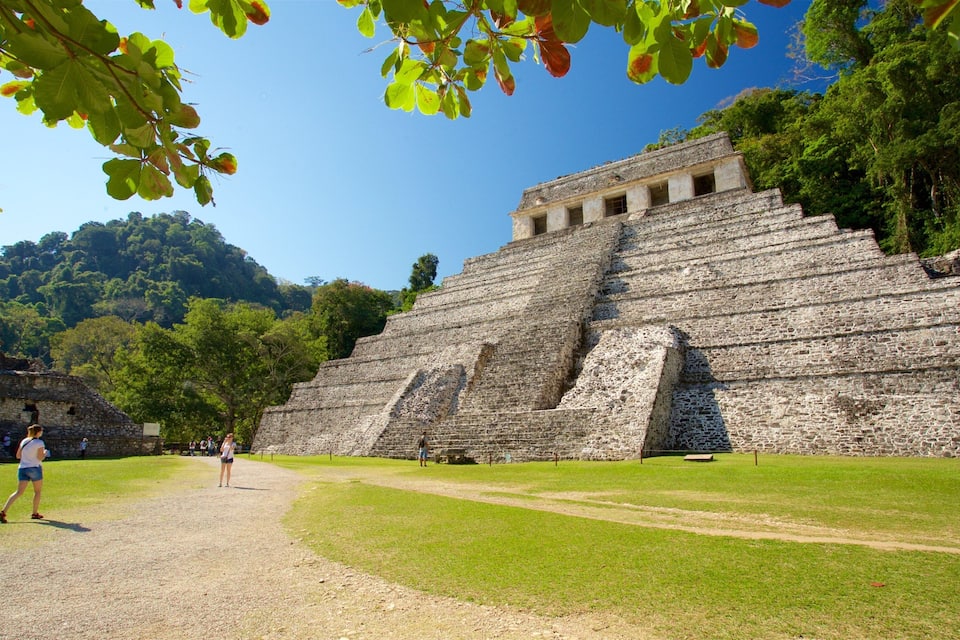 A large stone pyramid with a staircase leading to the top, surrounded by lush greenery and a clear blue sky.