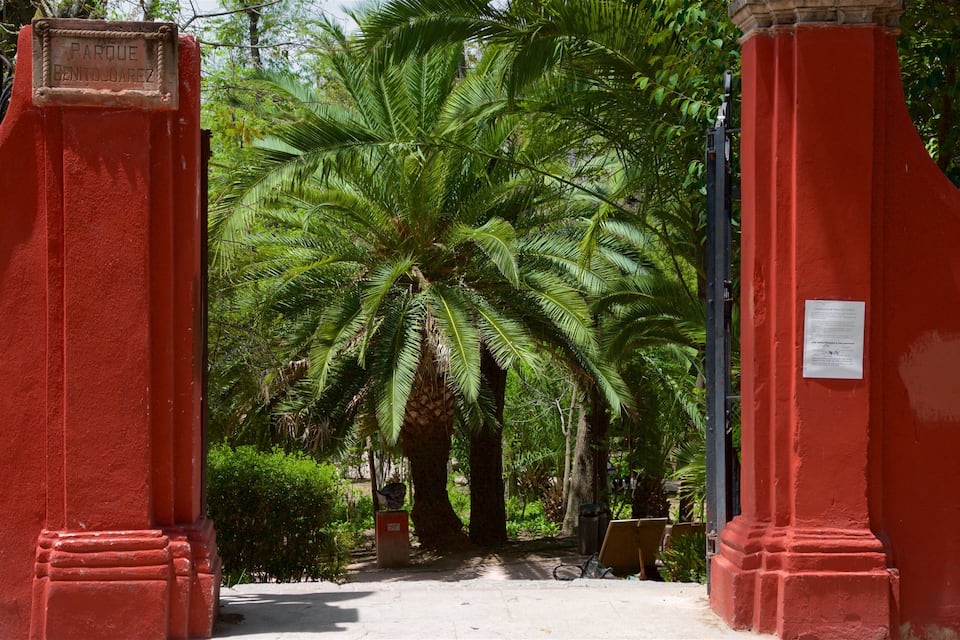 Een rode poort met een bord waarop 'Parque Benito Juarez' staat, die uitkomt op een pad omzoomd door palmbomen.