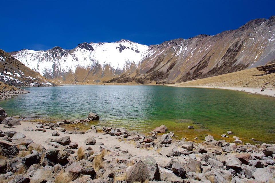 Um lago de montanha límpido, rodeado por terreno rochoso e picos cobertos de neve.