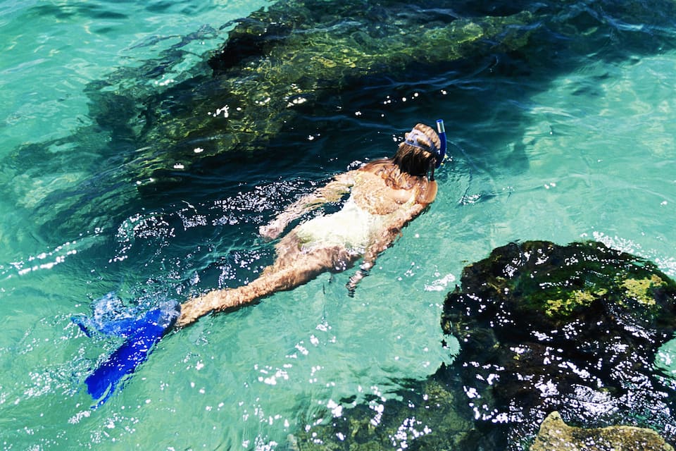 A person swimming in clear blue water with rocks.