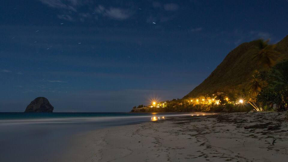 A beach at night with a mountain, a large rock, and lights on the hillside.