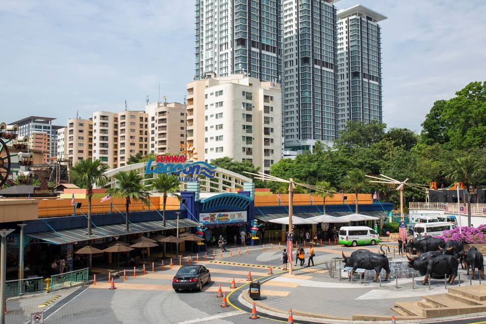A city street with a large building, palm trees, and a Ferris wheel.
