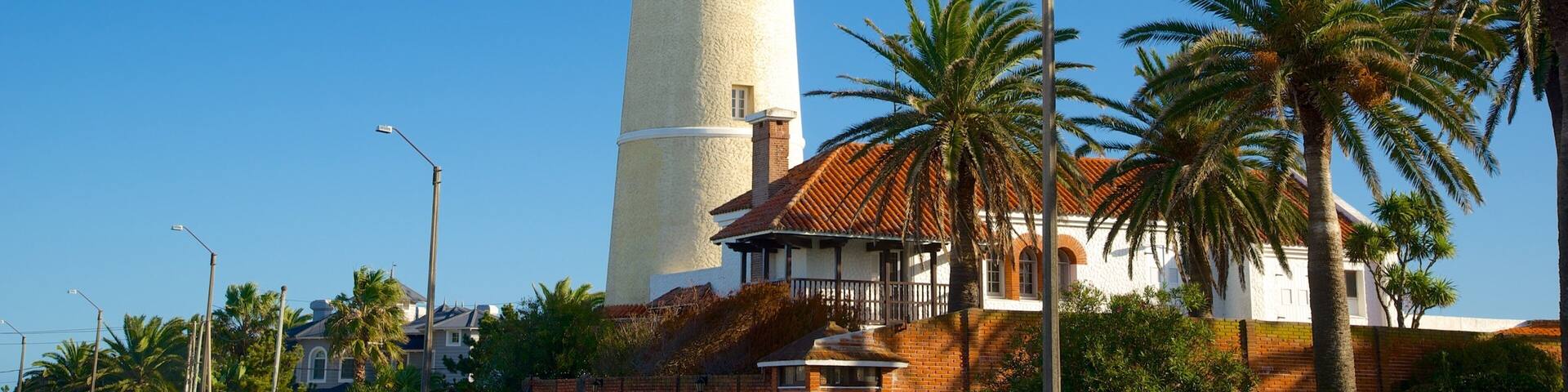 A tall white lighthouse with a red top, surrounded by palm trees and a building with a red-tiled roof.